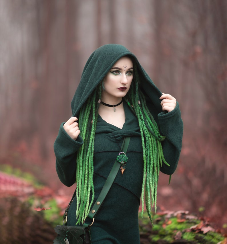 Peut inclure: Une femme portant une cape &agrave; capuche vert fonc&eacute; avec une longue tresse verte. Elle porte un collier avec un pendentif vert et une sangle en cuir. L'arri&egrave;re-plan est une for&ecirc;t floue.