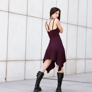May include: A woman wearing a burgundy asymmetrical dress with a criss-cross back detail and black knee-high boots. She is standing in front of a white tiled wall.