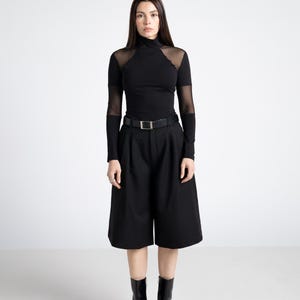 May include: A black outfit featuring a turtleneck top with sheer mesh accents on the shoulders and sleeves, paired with high-waisted culottes and a belt. The ensemble is completed with black pointed-toe ankle boots.