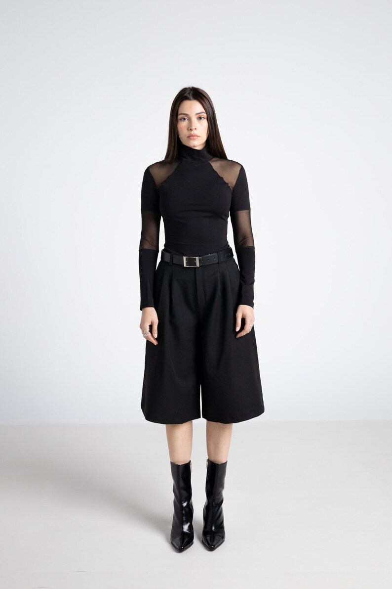 May include: A black outfit featuring a turtleneck top with sheer mesh accents on the shoulders and sleeves, paired with high-waisted culottes and a belt. The ensemble is completed with black pointed-toe ankle boots.
