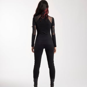 May include: Black long-sleeve top and pants. The top features sheer mesh panels on the shoulders and sleeves. The model is wearing black boots. The hair is dark with red highlights. The outfit is a sleek, modern style.
