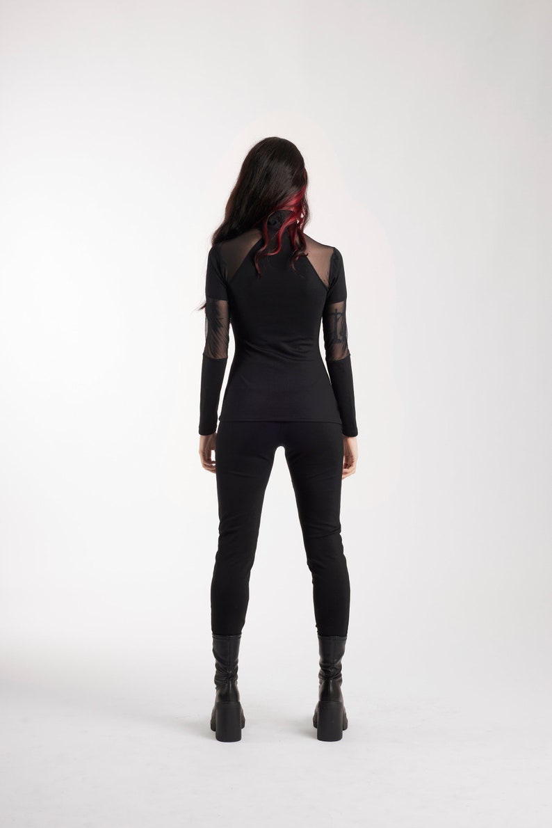 May include: Black long-sleeve top and pants. The top features sheer mesh panels on the shoulders and sleeves. The model is wearing black boots. The hair is dark with red highlights. The outfit is a sleek, modern style.