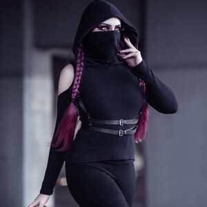 May include: A person wearing a black hooded top with a black face mask, black pants, and a black leather harness with buckles. The person has long, bright pink hair.