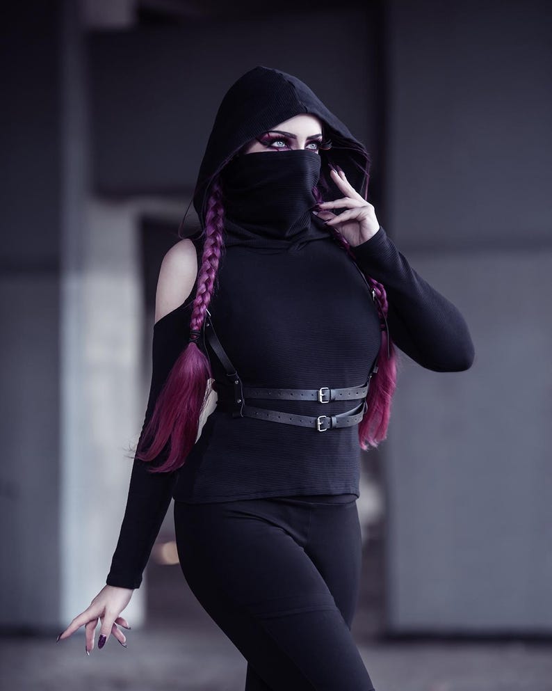 May include: A person wearing a black hooded top with a black face mask, black pants, and a black leather harness with buckles. The person has long, bright pink hair.