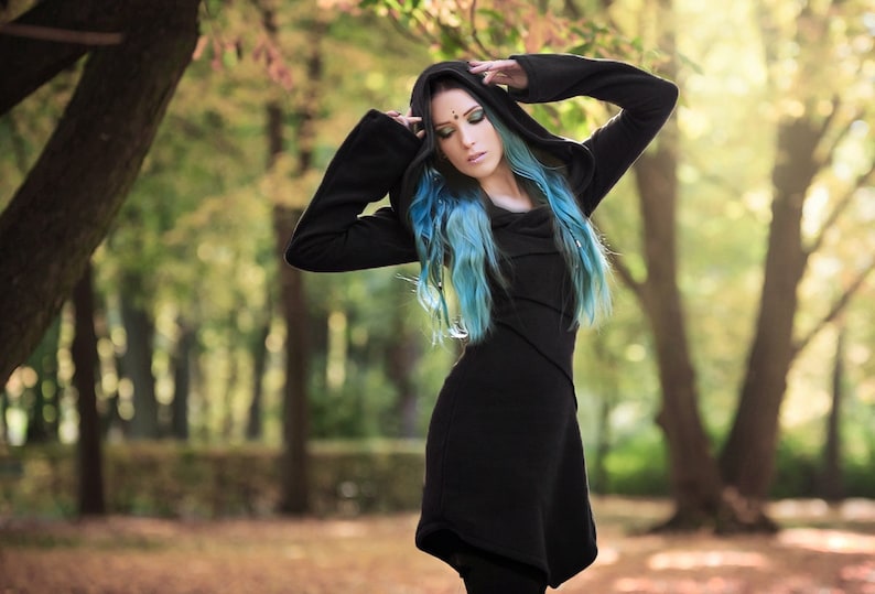 Peut inclure: Une femme aux cheveux bleus portant un sweat &agrave; capuche noir avec un long design fluide. Elle se tient dans une for&ecirc;t avec des arbres en arri&egrave;re-plan.