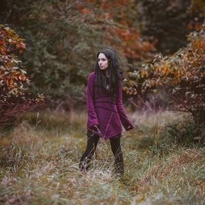 Peut inclure: Une femme portant une longue tunique &agrave; capuche de couleur pourpre fonc&eacute; avec une longue jupe noire et des bottes noires se tient dans un champ d'herbe haute. L'arri&egrave;re-plan est une image floue d'arbres avec des feuilles d'automne.