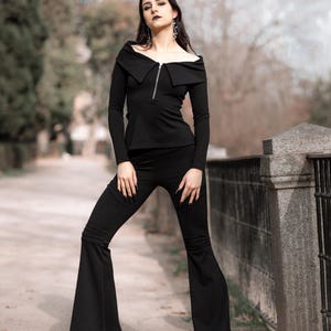 Puede incluir: Una mujer con un top negro de hombros descubiertos con cremallera y pantalones acampanados negros. Est&aacute; de pie frente a una valla met&aacute;lica negra y una pared de piedra.