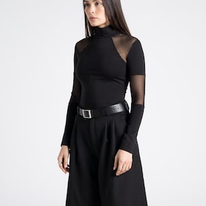 May include: A black outfit featuring a long-sleeved top with sheer mesh shoulder and upper arm panels, a high neckline, and black culottes. A black belt with a silver buckle cinches the waist. The outfit is a modern, minimalist style.