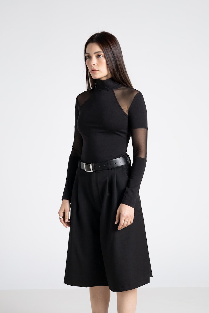 May include: A black outfit featuring a long-sleeved top with sheer mesh shoulder and upper arm panels, a high neckline, and black culottes. A black belt with a silver buckle cinches the waist. The outfit is a modern, minimalist style.
