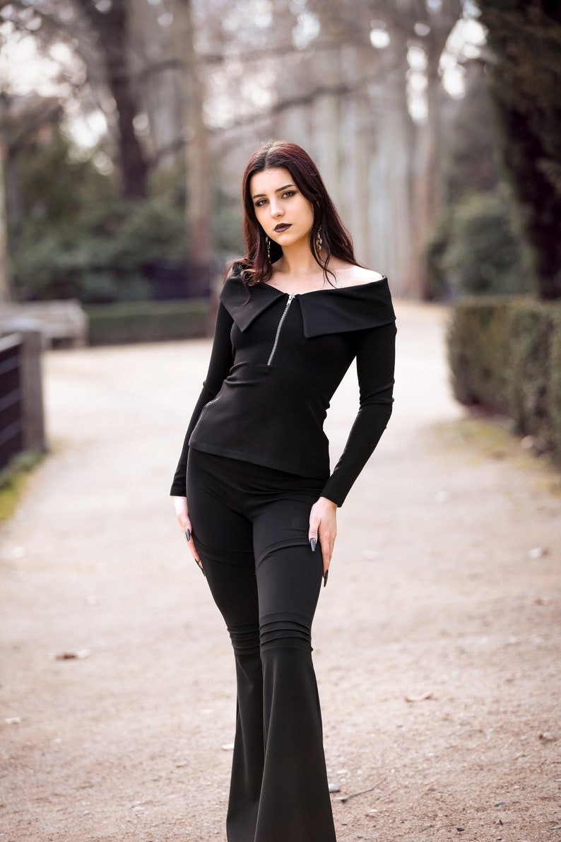 Puede incluir: Una mujer con un top negro de hombros descubiertos con cremallera y pantalones acampanados negros. Est&aacute; de pie en un camino en un parque.