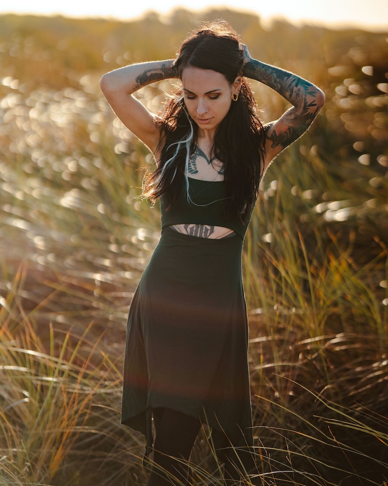 May include: A woman with long dark hair and tattoos stands in a field of tall grass. She is wearing a dark green dress with a cut-out detail at the waist. The sun is shining brightly, and the grass is a golden color.