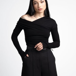 Op de afbeelding: Een zwart shirt met lange mouwen en een asymmetrische off-shoulder halslijn met een gekruist stofdetail over de borst. Het kledingstuk is nauwsluitend met lange mouwen en wordt gecombineerd met een zwarte broek met hoge taille en een dunne riem.