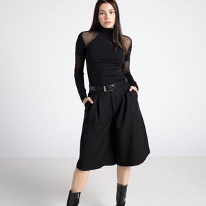 May include: A woman in a black outfit, including a turtleneck top with sheer mesh panels, high-waisted culottes, and pointed-toe ankle boots. A black belt cinches the waist. The outfit is all black, creating a sleek, modern look.