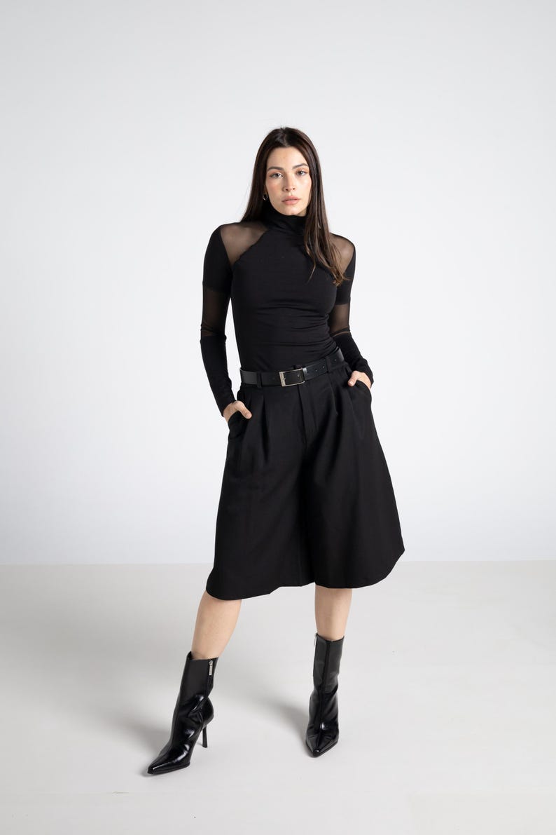 May include: A woman in a black outfit, including a turtleneck top with sheer mesh panels, high-waisted culottes, and pointed-toe ankle boots. A black belt cinches the waist. The outfit is all black, creating a sleek, modern look.
