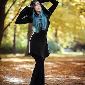 Peut inclure: Une femme aux cheveux bleus porte une tunique noire &agrave; capuche et un pantalon noir. Elle se tient dans une for&ecirc;t avec des feuilles mortes au sol.