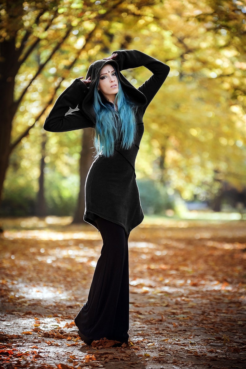 Peut inclure: Une femme aux cheveux bleus porte une tunique noire &agrave; capuche et un pantalon noir. Elle se tient dans une for&ecirc;t avec des feuilles mortes au sol.