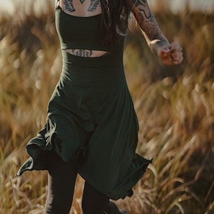 May include: A woman wearing a dark green dress with a cut-out detail at the waist, running through a field of tall grass.