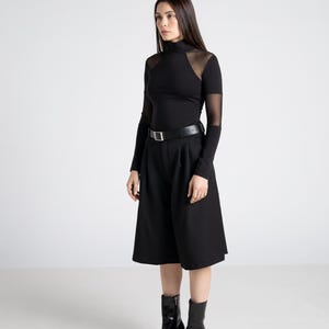 May include: A woman in a black outfit, featuring a long-sleeved top with sheer mesh panels at the shoulders and sleeves, paired with high-waisted culottes and a black belt. She wears black heeled ankle boots. The background is plain white.