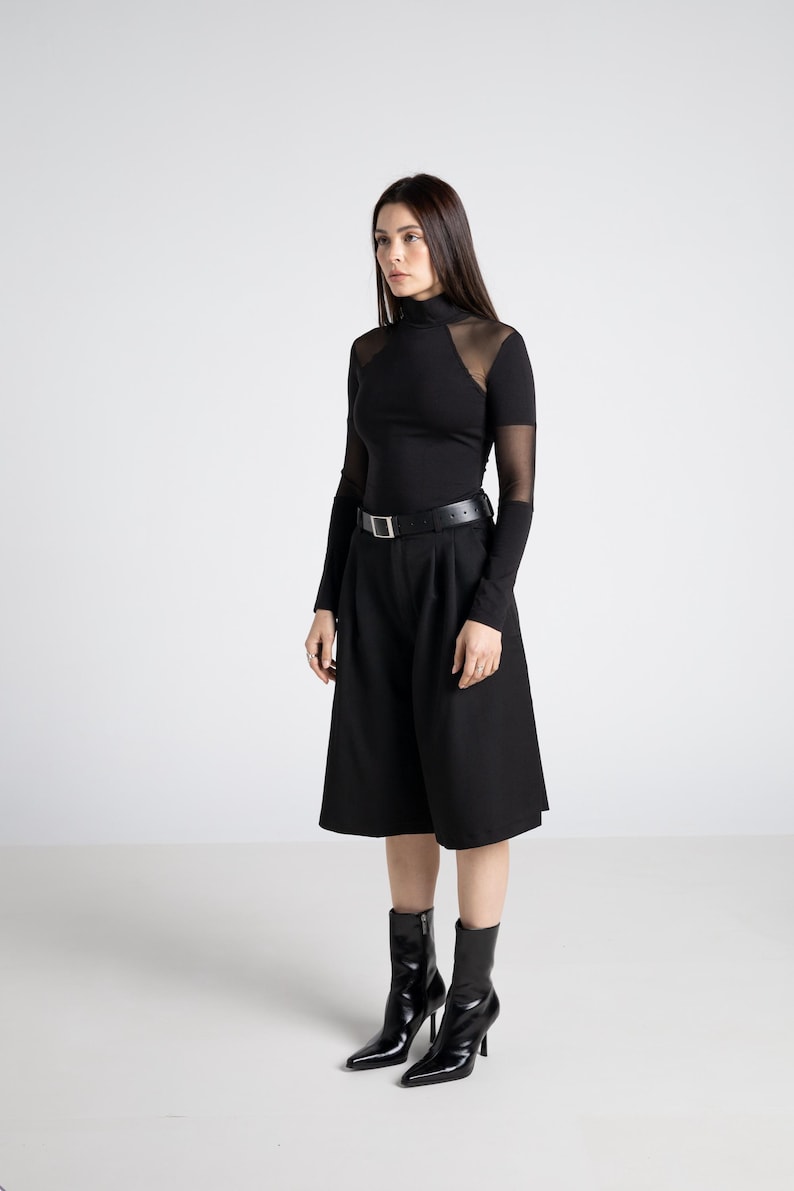 May include: A woman in a black outfit, featuring a long-sleeved top with sheer mesh panels at the shoulders and sleeves, paired with high-waisted culottes and a black belt. She wears black heeled ankle boots. The background is plain white.