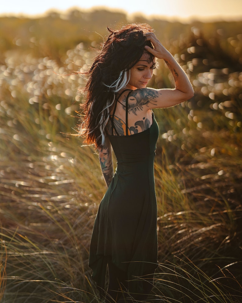 May include: A woman with long dark hair and tattoos wears a dark green dress in a field of tall grass.