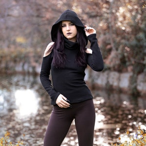 May include: A woman wearing a black hooded top with cut-out shoulders and brown flared pants. She is standing in a wooded area with a body of water in the background.