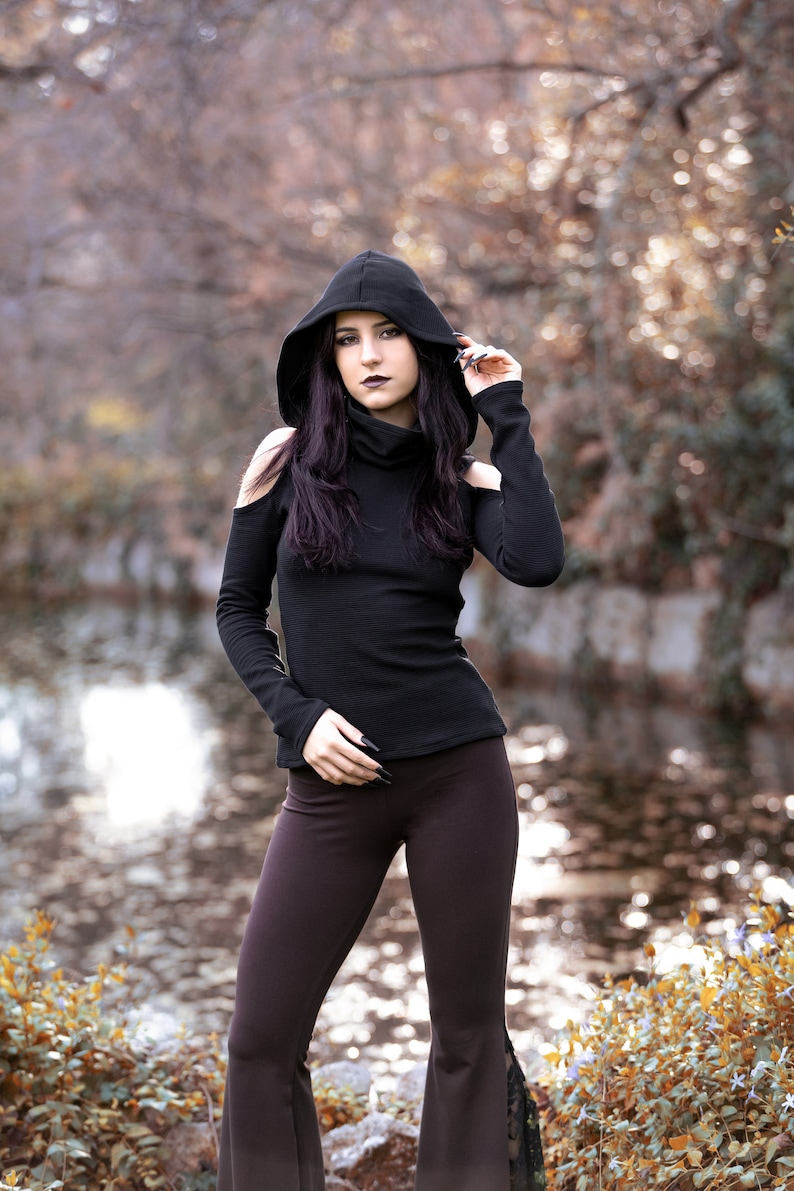 May include: A woman wearing a black hooded top with cut-out shoulders and brown flared pants. She is standing in a wooded area with a body of water in the background.