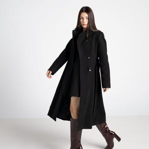 Puede incluir: Un abrigo largo negro de lana con cierre cruzado y cinturón en la cintura. El abrigo se combina con un vestido negro y botas altas burdeos con tacones altos. El abrigo tiene un diseño clásico y entallado.