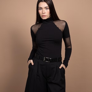 May include: Black turtleneck top with sheer mesh shoulder and sleeve accents, paired with black culottes and a black belt. The outfit is a monochromatic ensemble, creating a sleek and modern look. The model is wearing the outfit.