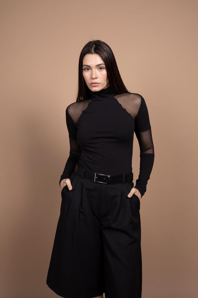 May include: Black turtleneck top with sheer mesh shoulder and sleeve accents, paired with black culottes and a black belt. The outfit is a monochromatic ensemble, creating a sleek and modern look. The model is wearing the outfit.