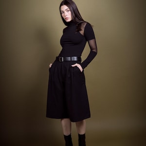 May include: A woman in a black outfit, featuring a turtleneck top with sheer sleeves, a belted skirt, and ankle boots. The outfit is all black, creating a sleek and modern look. The background is a solid olive green.