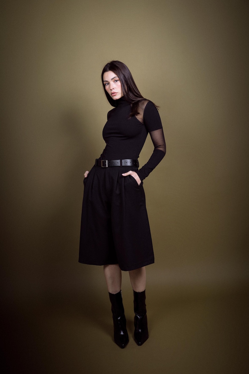 May include: A woman in a black outfit, featuring a turtleneck top with sheer sleeves, a belted skirt, and ankle boots. The outfit is all black, creating a sleek and modern look. The background is a solid olive green.