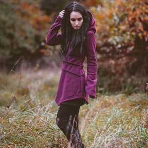 Peut inclure: Une femme portant un long sweat &agrave; capuche violet avec un legging noir. Elle se tient dans un champ d'herbe haute.