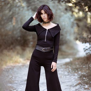 Puede incluir: Una mujer con un mono negro de pierna ancha. El mono tiene una cremallera y un cuello ancho. Lleva un cintur&oacute;n negro con una hebilla plateada. La mujer tiene el pelo oscuro y est&aacute; de pie en un entorno de bosque.