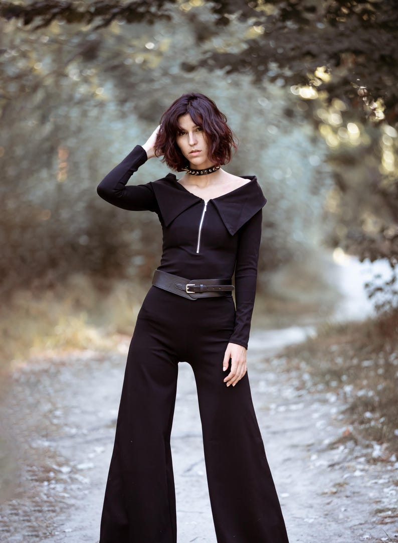 Puede incluir: Una mujer con un mono negro de pierna ancha. El mono tiene una cremallera y un cuello ancho. Lleva un cintur&oacute;n negro con una hebilla plateada. La mujer tiene el pelo oscuro y est&aacute; de pie en un entorno de bosque.