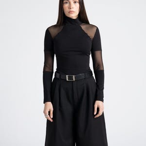 May include: A black turtleneck top with sheer mesh accents on the shoulders and sleeves, paired with black culottes and a black belt with a silver buckle. The outfit is a monochromatic ensemble.