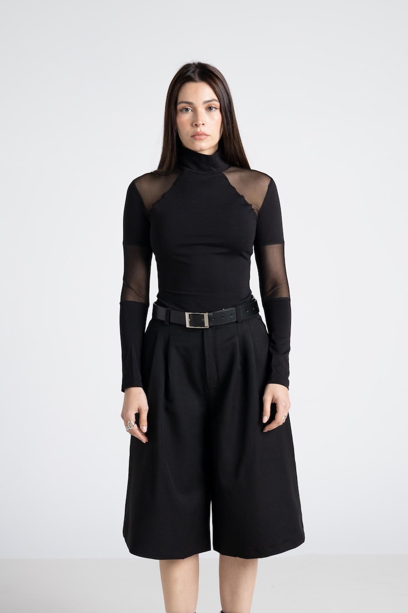 May include: A black turtleneck top with sheer mesh accents on the shoulders and sleeves, paired with black culottes and a black belt with a silver buckle. The outfit is a monochromatic ensemble.