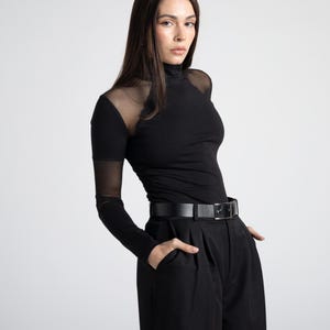 May include: Black turtleneck top with sheer mesh sleeves and shoulders, paired with black high-waisted pants and a black belt. The outfit is a monochromatic ensemble, creating a sleek and modern look. The pants have a pleated design.
