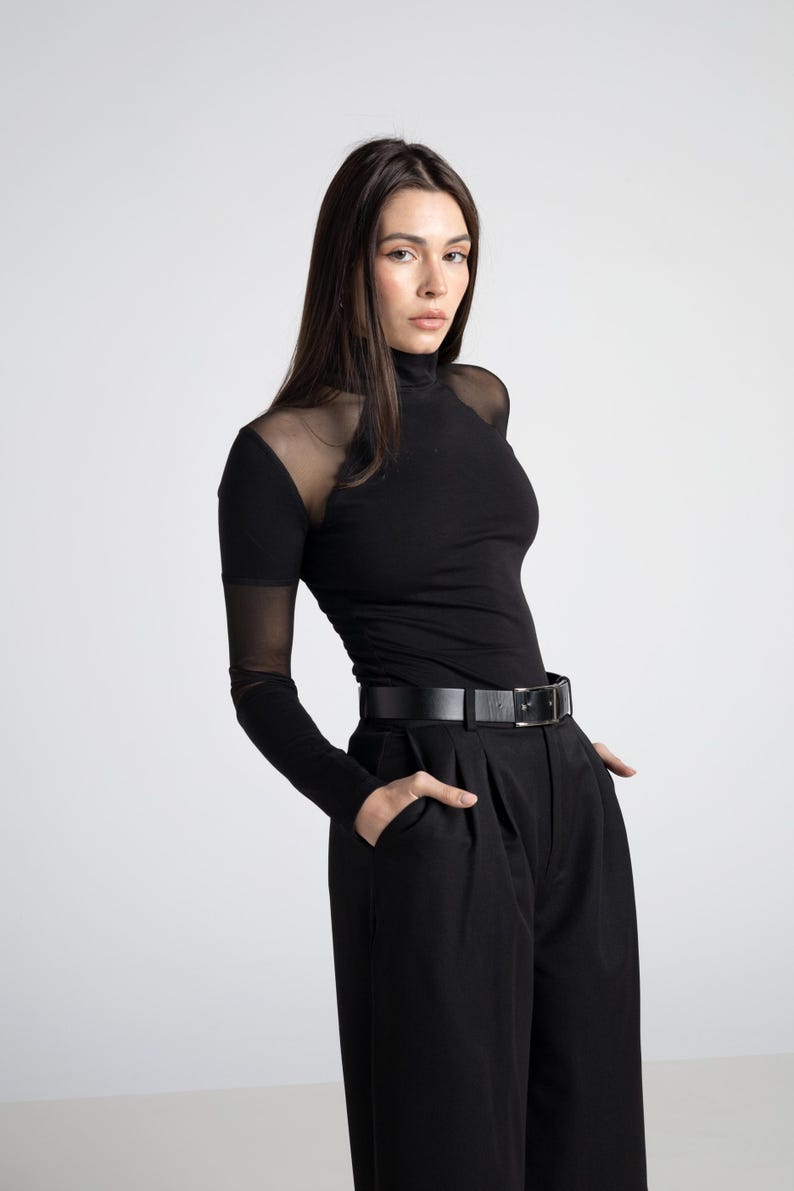 May include: Black turtleneck top with sheer mesh sleeves and shoulders, paired with black high-waisted pants and a black belt. The outfit is a monochromatic ensemble, creating a sleek and modern look. The pants have a pleated design.