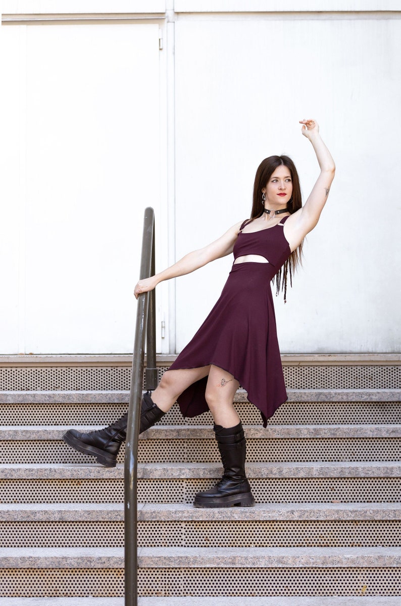 May include: A woman wearing a burgundy asymmetrical dress with a cut-out detail, black boots, and a black choker necklace. She is standing on a set of stairs with a metal railing.