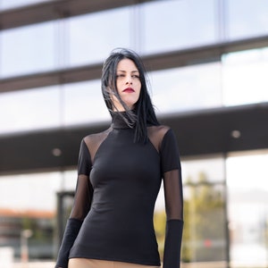 May include: A black long-sleeved top with sheer mesh panels on the shoulders and sleeves. The top has a high neckline and is paired with tan-colored pants. The outfit is worn outdoors in front of a modern building.