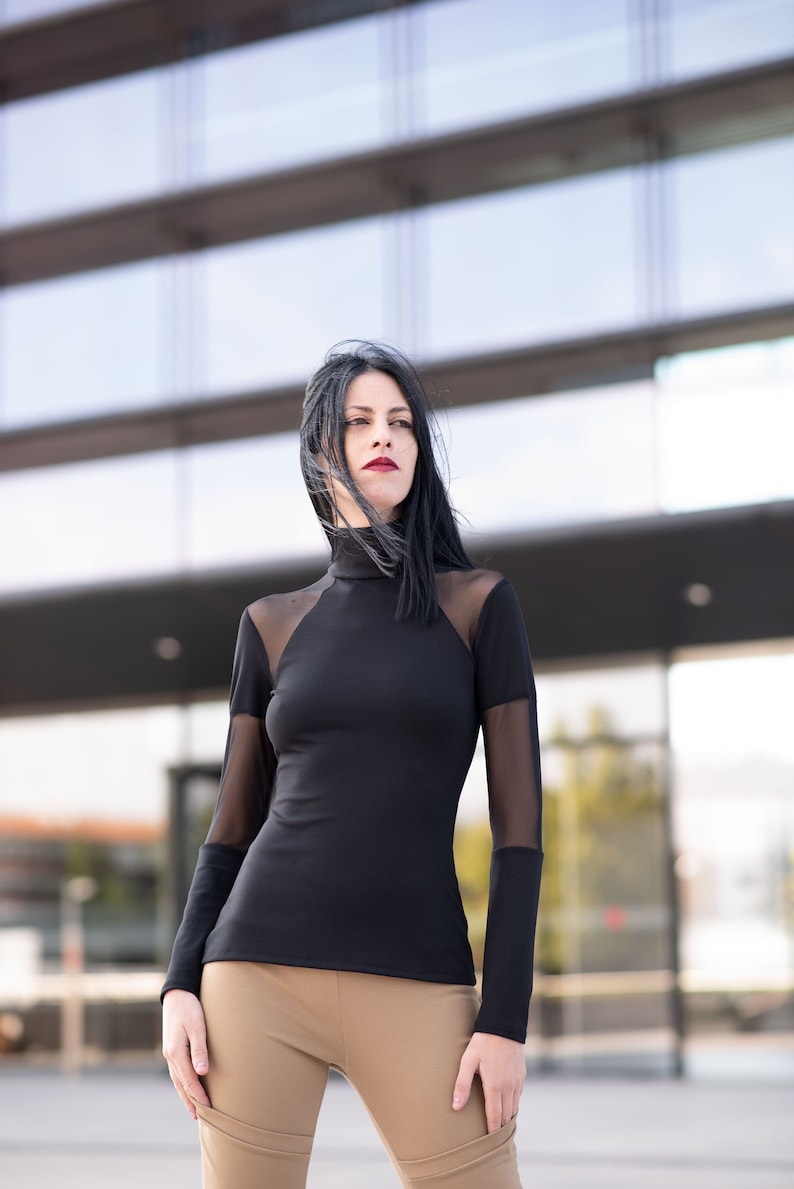 May include: A black long-sleeved top with sheer mesh panels on the shoulders and sleeves. The top has a high neckline and is paired with tan-colored pants. The outfit is worn outdoors in front of a modern building.