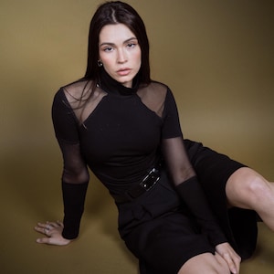 May include: A black top with sheer mesh panels on the shoulders and sleeves, paired with a matching black skirt and belt. The outfit is worn by a person sitting on a gold-toned surface. The top has a high neckline.