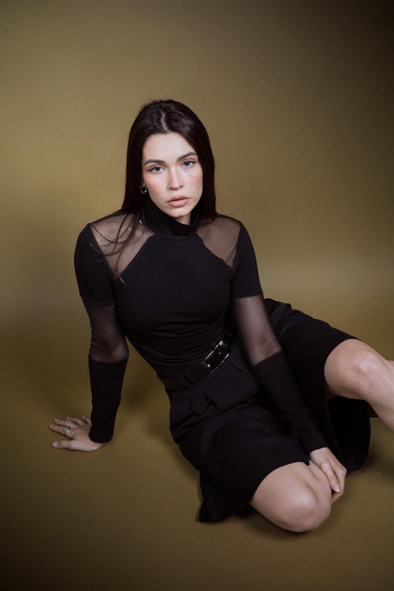 May include: A black top with sheer mesh panels on the shoulders and sleeves, paired with a matching black skirt and belt. The outfit is worn by a person sitting on a gold-toned surface. The top has a high neckline.