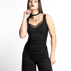 May include: A woman wearing a black sleeveless top with a sheer mesh panel and a black choker necklace. She is wearing black wide-leg pants.