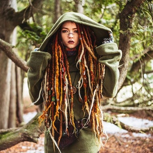 Peut inclure: Une femme avec de longues dreadlocks color&eacute;es porte un sweat-shirt &agrave; capuche vert avec une ceinture en cuir marron. Elle se tient dans un environnement forestier.