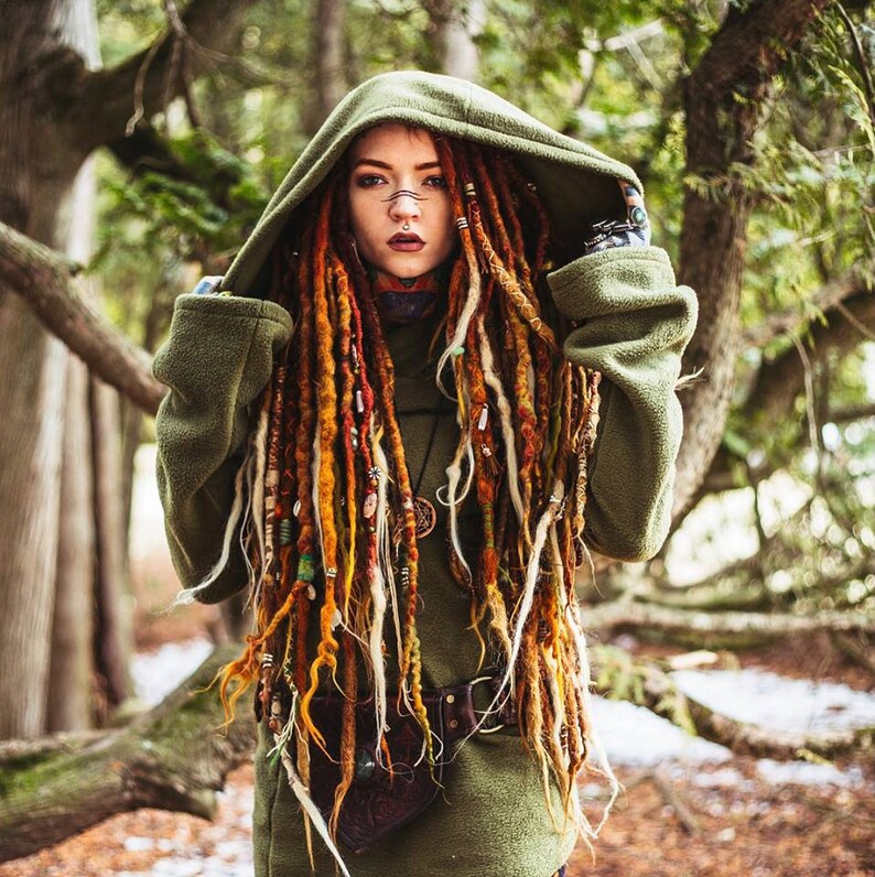 Peut inclure: Une femme avec de longues dreadlocks color&eacute;es porte un sweat-shirt &agrave; capuche vert avec une ceinture en cuir marron. Elle se tient dans un environnement forestier.