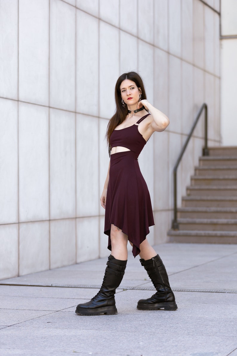 May include: A woman wearing a burgundy dress with a cut-out detail and black knee-high boots. She is standing in front of a white tiled wall.