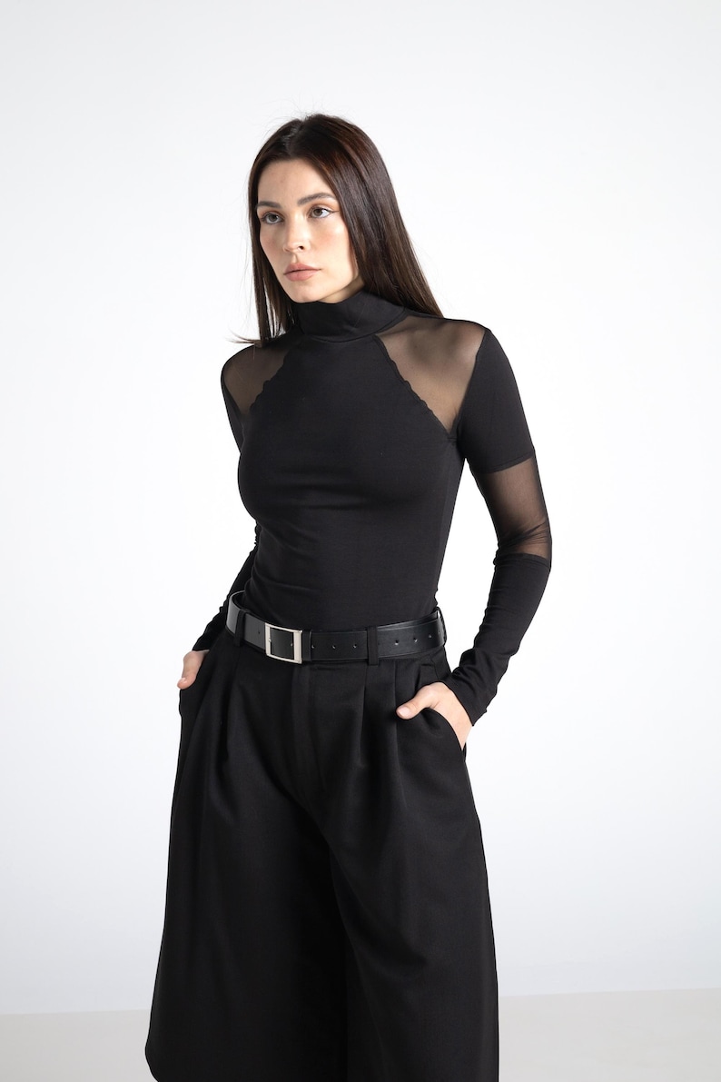 May include: Black long-sleeved top with a high neckline and sheer mesh shoulder and sleeve accents. The top is paired with black wide-leg pants and a black belt with a silver buckle. The outfit is a monochromatic ensemble.