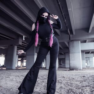 May include: A woman wearing a black hooded top, black pants, and a black face mask. She has pink hair and is posing with her hand in a peace sign. The photo is taken in a concrete underpass.