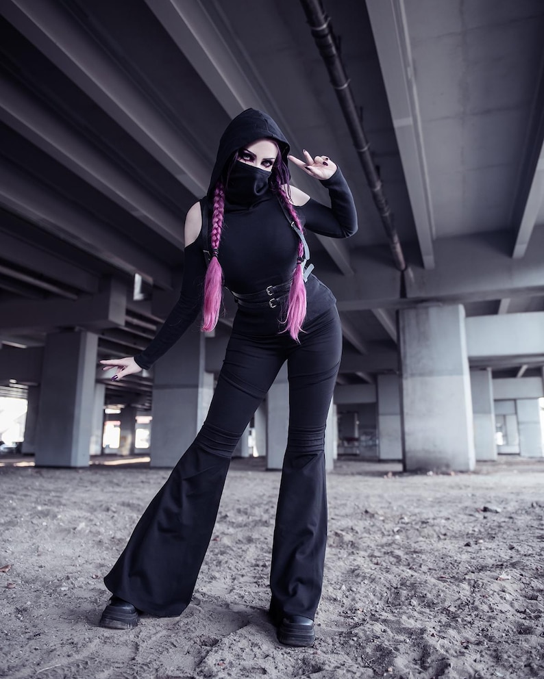 May include: A woman wearing a black hooded top, black pants, and a black face mask. She has pink hair and is posing with her hand in a peace sign. The photo is taken in a concrete underpass.